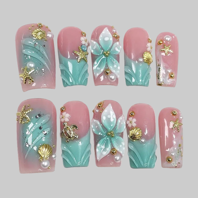 Aqua Bloom évoque la rencontre entre l’océan et la fleur. Sur une base rose poudré et turquoise nacré, des pétales sculptés, des perles et des éléments dorés — étoiles de mer, coquillages, tortue — créent une manucure à la fois élégante et lumineuse.
Ce modèle incarne la fraîcheur marine et la féminité délicate, parfait pour un look estival raffiné. press-on nails faux ongles bleu et rose