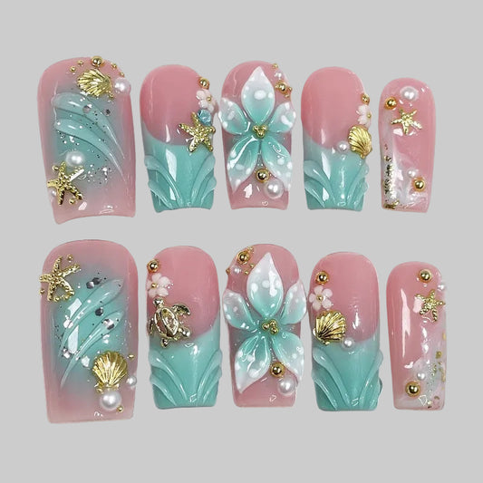 Aqua Bloom évoque la rencontre entre l’océan et la fleur. Sur une base rose poudré et turquoise nacré, des pétales sculptés, des perles et des éléments dorés — étoiles de mer, coquillages, tortue — créent une manucure à la fois élégante et lumineuse.
Ce modèle incarne la fraîcheur marine et la féminité délicate, parfait pour un look estival raffiné. press-on nails faux ongles bleu et rose