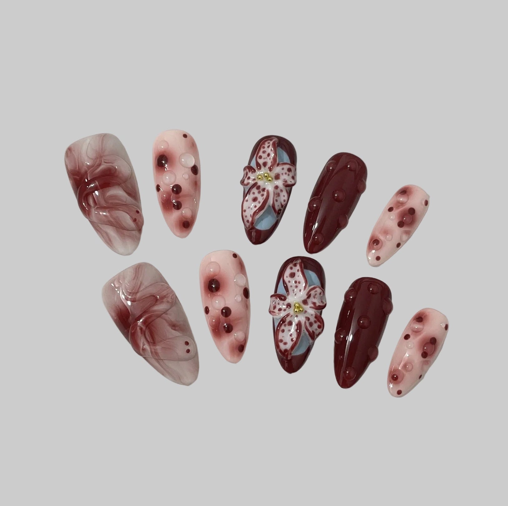 Burgundy Orchid incarne la sophistication et la passion. Sur une base rose poudré et bordeaux profond, des fleurs sculptées en relief aux pétales tachetés se mêlent à des effets marbrés et des perles délicates pour une manucure à la fois artistique et sensuelle.
Ce modèle évoque la beauté d’une orchidée rare — élégante, mystérieuse et pleine de caractère.  rouge bordeau, été, summer ongles press-on nails 
